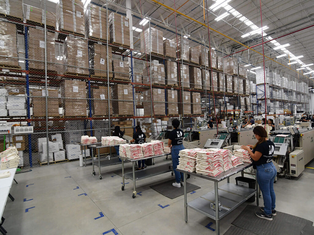 Workers in a large factory sort and package stacks of goods, including Monster Digital apparel printing products, on tables. Tall shelves full of boxes line the organized warehouse, known for its quality processes and bright, industrial atmosphere.