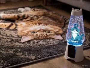A realistic cat rug with a cat’s face and paws lies on the floor. In the foreground, a blue lava lamp glows, featuring a cartoon cat inside—making these funny cat gifts perfect for anyone searching for weird gifts for cat lovers.