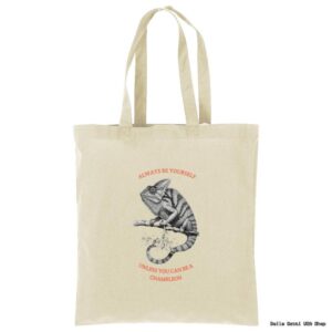 A beige canvas tote bag features a black and white sketch of a chameleon on a branch. Above and below the chameleon, red text reads: ALWAYS BE YOURSELF UNLESS YOU CAN BE A CHAMELEON. The bag has two handles.