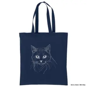 A navy blue tote bag featuring a minimalist white line drawing of a cats face with expressive eyes and detailed fur. The bag has two long handles for carrying and a plain background, highlighting the simple yet elegant cat illustration.