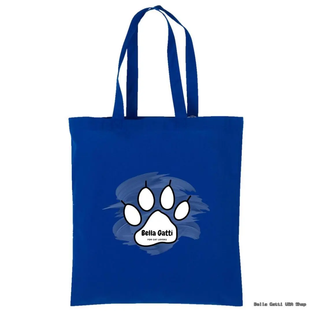 A blue tote bag features a white paw print graphic in the center, outlined in black, with “Bella Gatti” and “For Cat Lovers” written inside the paw. Behind the paw is a painterly light blue brushstroke background. The bag has two handles.
