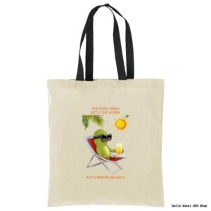A beige tote bag with black handles features an illustration of a relaxed green bird wearing sunglasses, lounging on a deck chair with a drink. Above it reads, “THE EARLY BIRD GETS THE WORM,” and below, “BUT I PREFER BRUNCH!” A sun shines overhead.