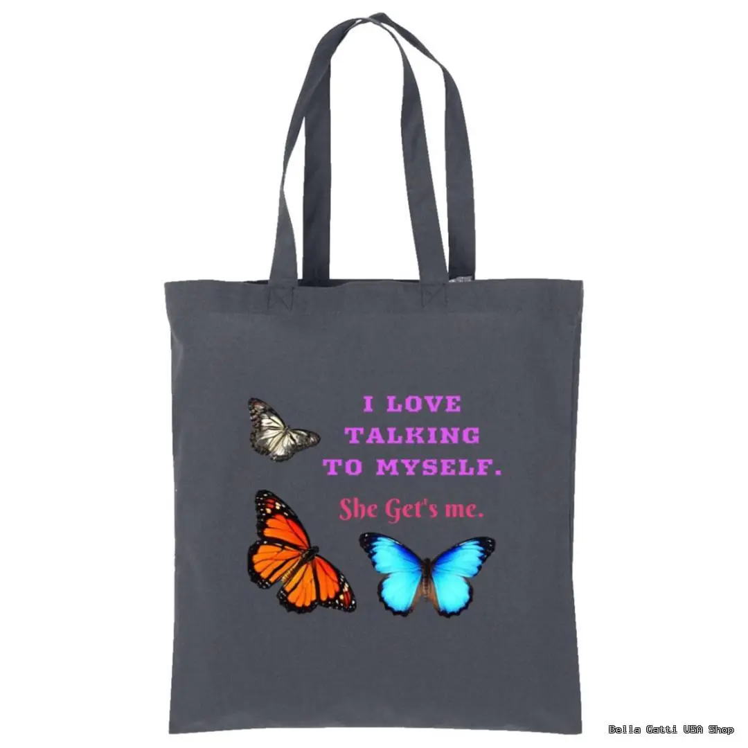 A dark gray tote bag with two handles, decorated with three butterflies—one monarch, one blue, and one gray. The text in bold pink and white reads: “I LOVE TALKING TO MYSELF. She Get’s me.” The text is centered between the butterflies.