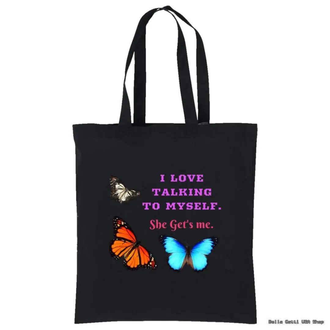 A black tote bag with two butterflies, one orange and black, one blue. White and pink text reads: I LOVE TALKING TO MYSELF. She Gets me. The design is centered and vibrant, with the butterflies flanking the text.
