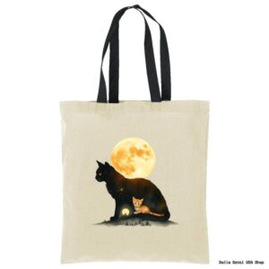 A beige tote bag with black handles features artwork of a large black cat silhouette with a glowing full moon inside it. A small orange cat lies in front of the silhouette, which also contains trees and a house, creating a dreamy, magical scene.