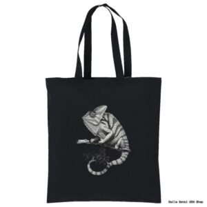 A black tote bag features a detailed grayscale illustration of a chameleon perched on a branch. The chameleon is facing left, and its curled tail and textured skin stand out against the dark background.