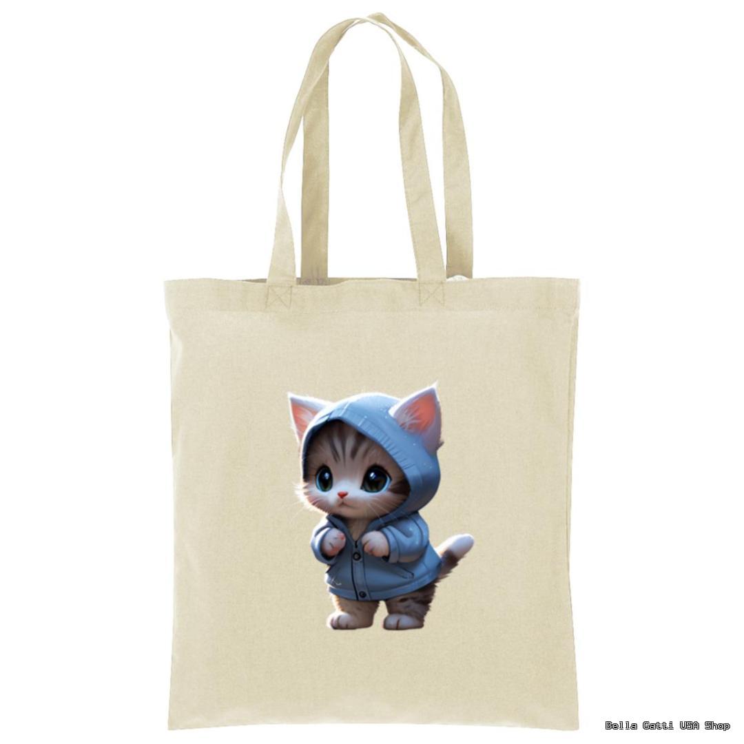 A beige tote bag featuring a printed illustration of a cute, wide-eyed kitten wearing a blue hoodie, standing upright on its hind legs with its front paws raised as if adjusting the hood. The background is plain and the design is centered.